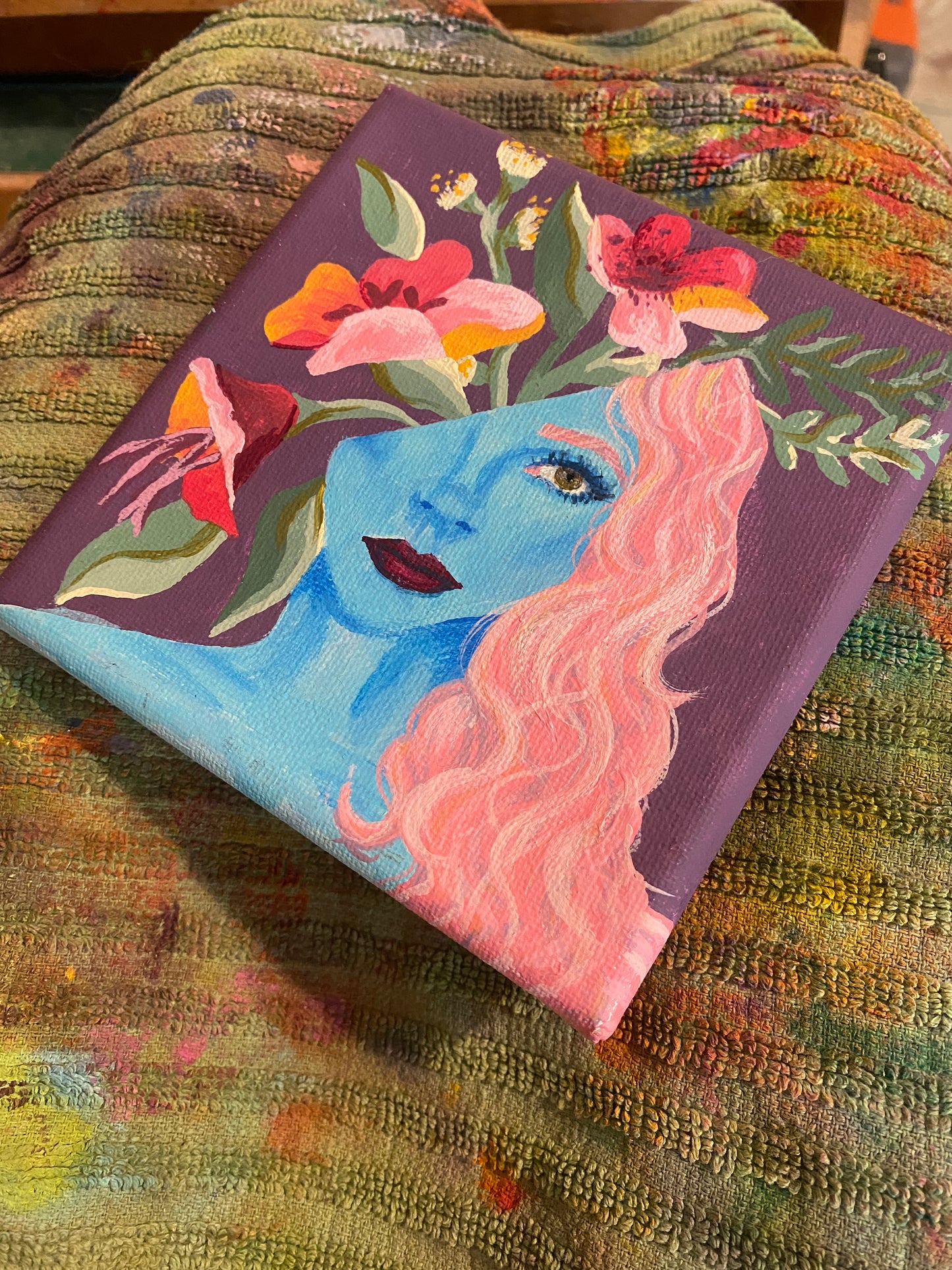 painting of a woman with blue skin and pink hair with her head split and flowers coming out on a paint towel