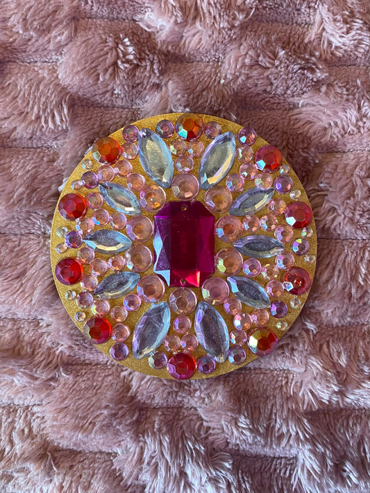 round magnet painted gold with pink and red small gemstones around the edges. White marquise cut gems surrounding a hot pink emerald cut stone in the center. 